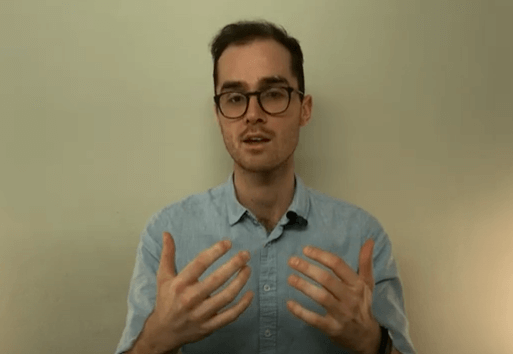 Man in glasses and blue shirt discussing acupuncture with hands raised against a plain background.