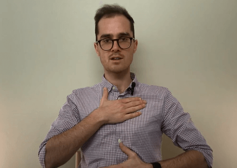 Man in glasses demonstrating tcm breathing technique against a plain background.