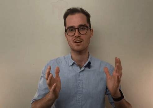 A man in glasses discusses acupuncture, gesturing with his hands while speaking indoors.
