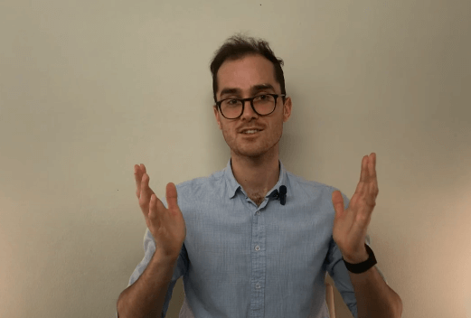 Man wearing glasses gestures with both hands, discussing acupuncture, in front of a plain wall.