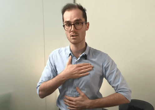 A man in glasses demonstrates a tcm technique, touching his chest and abdomen with his hands.