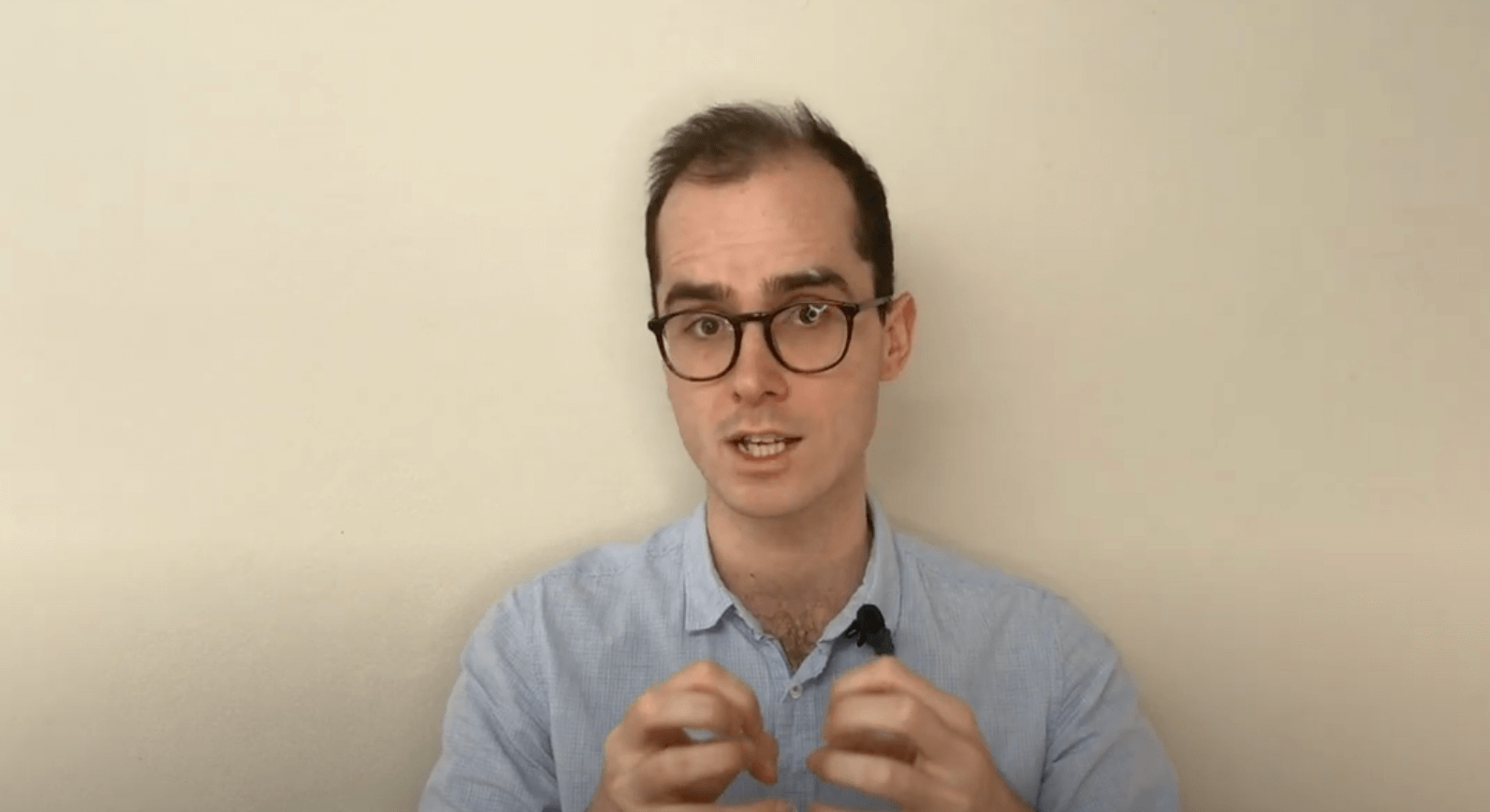 Man with glasses and blue shirt explains acupuncture while gesturing against a plain background.