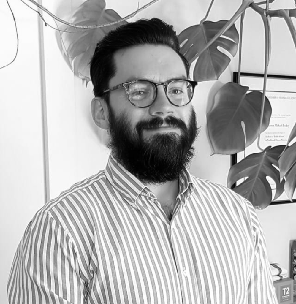Man with glasses and beard in striped shirt stands indoors near plant, framed TCM certificate.