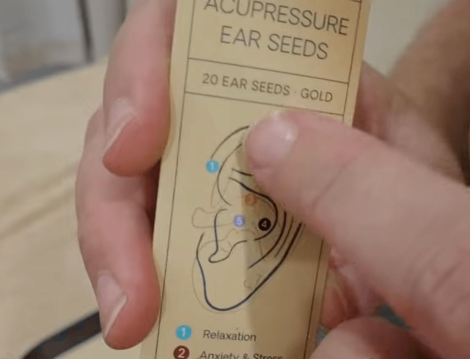Hand holding a pack of acupuncture ear seeds with diagram and labeled pressure points.